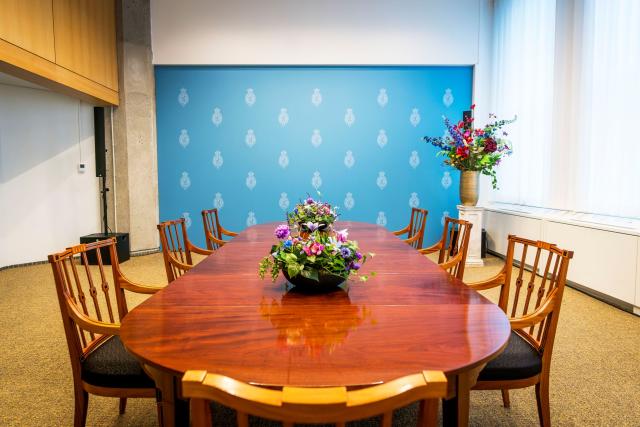 A room with a blue wall in which a large wooden table is placed. Seven chairs are set around it and flowers are placed on the table as centre pieces.