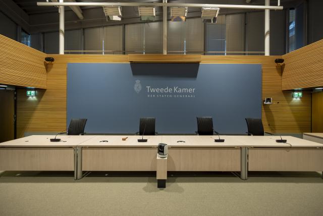 A room specifically designed for inquiries. Four chairs are placed behind a long table. Behind the setup is a blue screen with the "Tweede Kamer" logo.