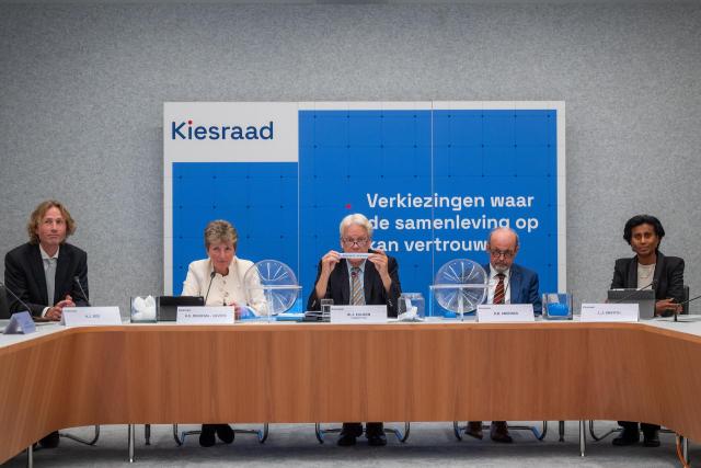 The electoral Council sits behind a panel in front of a blue screen. One of the members is holding up a slip of paper which covers a part of his face.