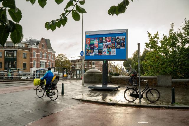 A billboard with campaign posters is set up on the sidewalk. A cyclist cycles by. Across the street there are several houses and on the right hand side of the bill board someone has parked a bike.