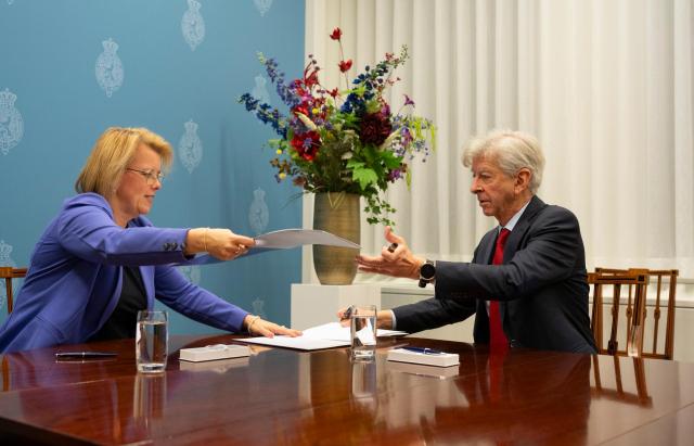 The temporary chairman and the informateur are seated at the official "schrijftafel" which forms the scene of the formation. The chairman is handing over a document to the informateur, who is reaching out to receive it.